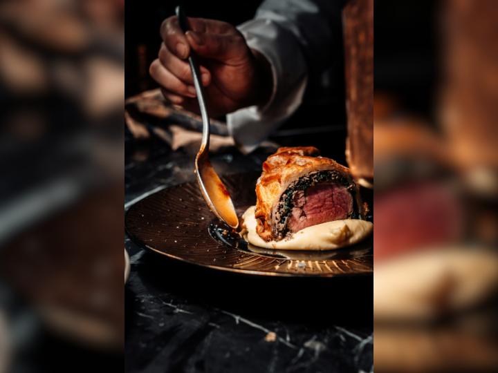 A close up image of a piece of steak being plated. A Black hand is holding a spoon that is dipping into the sauce next to the steak.