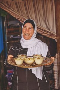 A brown-skinned woman in a white headscarf holds up a tray of rice pudding.
