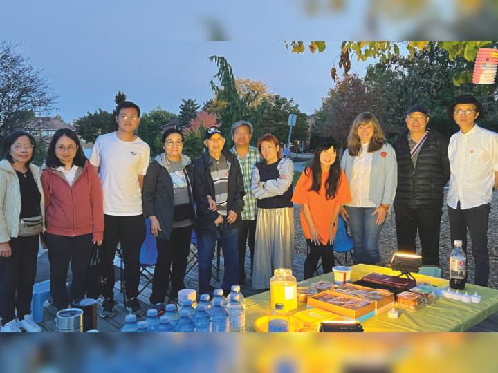 Photo of 11 people standing in front of a table with snacks and bottled water. Ten of them are East Asian and one woman is a middle-aged white woman. The group of people vary in age from young adults to middle-aged people.