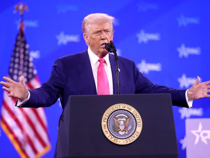 Donald Trump speaking at a podium with arms outstretched. Blue CPAC backdrop and American flag behind him.